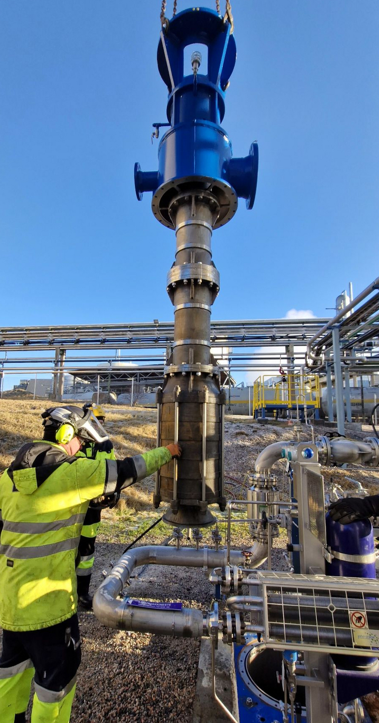 A vertical high-pressure pump (Apollotyp GLKV, VS1 according to API 610) hangs from a crane and is inserted into the pump can. The pump is installed in a liquid ammonia plant while a worker in safety clothing supervises the crane assembly. An industrial plant and blue, cloudless sky can be seen in the background.