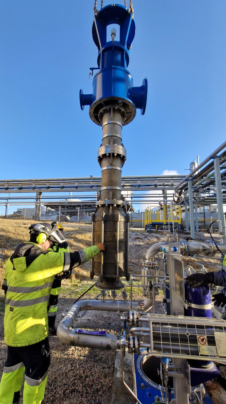 Eine vertikale Hochdruckpumpe (Apollotyp GLKV, VS1 gemäß API 610) hängt an einem Kran und wird in den Pumpentopf eingesetzt. Die Pumpe wird in einer Flüssigammoniak Anlage montiert während ein Arbeiter in Sicherheitskleidung die Kranmontage beaufsichtigt. Im Hintergrund ist eine Industrieanlage und blauer, wolkenloser Himmel zu sehen.