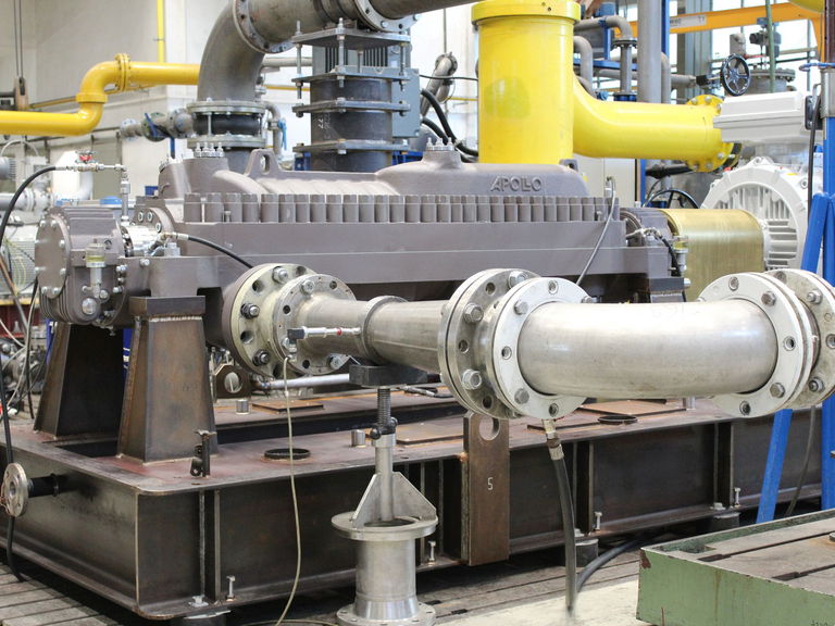 Das Hochdruckpumpenaggregat BB3 ist auf dem Apollo-Prüfstand an einer Rohrleitung montiert. Im Hintergrund sind weitere Verrohrungen des Testfelds zu sehen. Der Grundrahmen des Pumpenaggregats besteht aus Rohstahl, die Pumpe aus Gusseisen ist noch nicht lackiert.