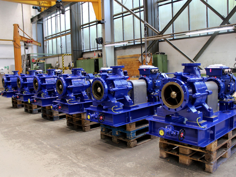 Zu sehen ist eine leicht schräg von vorne aufgenommene Gruppe von mehrstufigen GLZ-Prozesskreiselpumpen, die in einer Reihe auf Holzpaletten hintereinander in der Fertigungshalle von Apollo stehen. Die Pumpen werden kurz vor der Endabnahme fotografiert. 