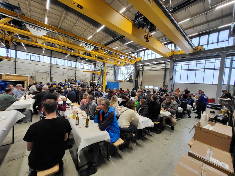 Überblicksbild in die Fertigungshalle von Apollo Gößnitz, in der alle Mitarbeiter und Mitarbeiterinnen an Bierzeltgarnituren sitzen und der Geschäftsführung bei der Rede zuhören.