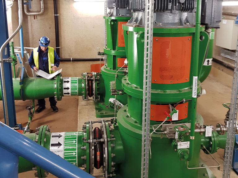 A service technician standing by a pipeline in a power station hall with green vertical process pumps, checking the papers