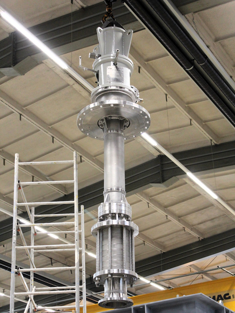 A VS1 pump hangs from the indoor crane. Next to it stands a metal scaffold. The indoor roof and lamps can be seen in the background.
