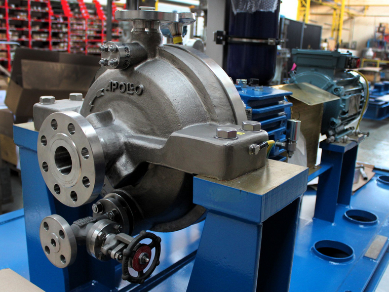 Detailed close-up of an OH2 API 610 pump with blue base frame and bearing bracket. The Apollo is cast into the pump housing. A red storage rack for screws and workshop equipment can be seen in the background.