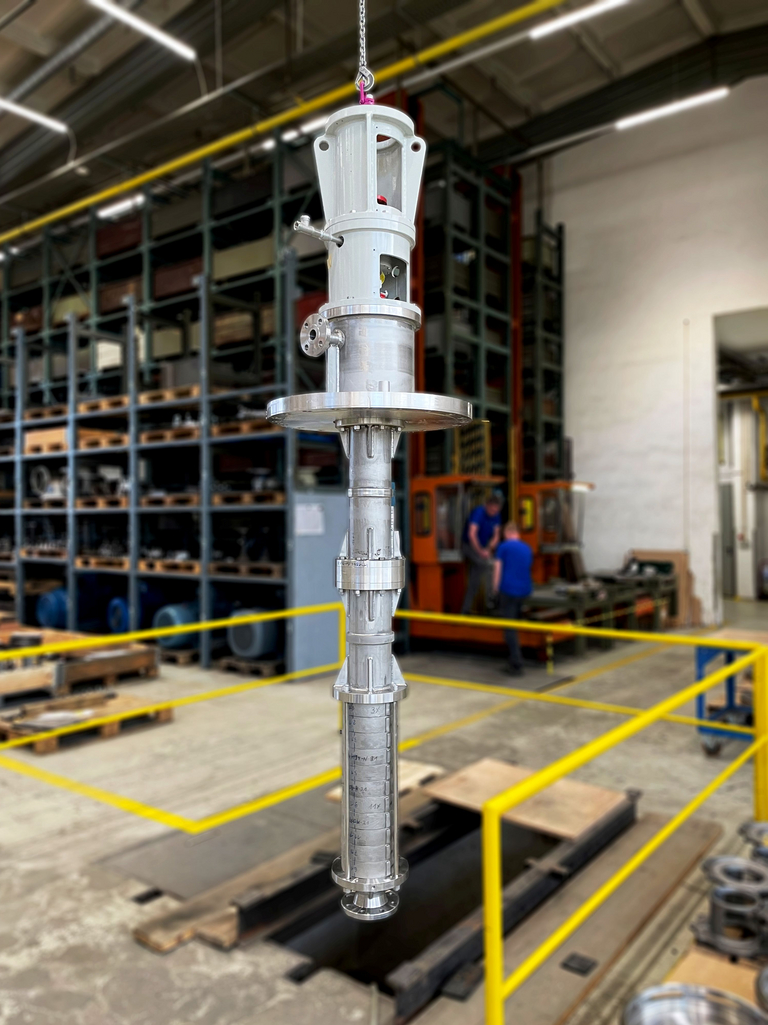 A GLV (VS1, API 610 pump) is suspended from the overhead crane. Below the vertical pump, there is an open assembly pit, which is enclosed by yellow railings for safety. In the background, a high-bay warehouse is visible.