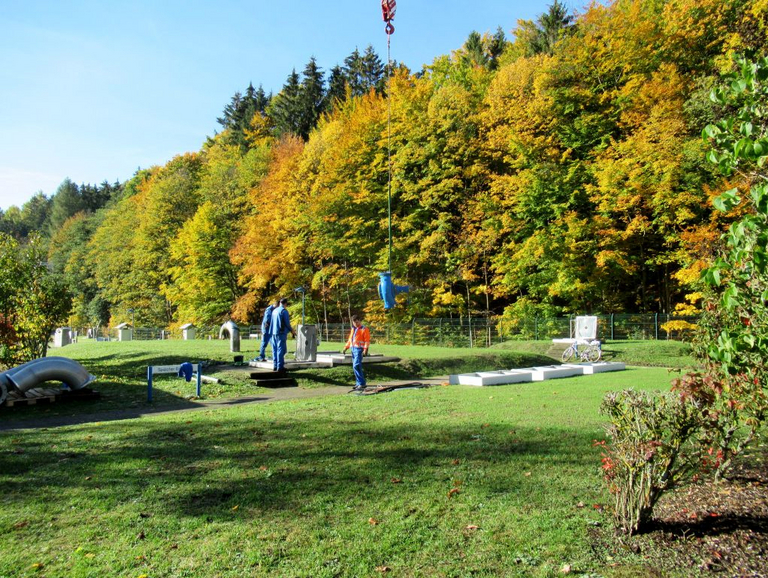 Zu sehen ist eine grüne Wiese mit einem Hügel in der Mitte. Im Hintergrund befindet sich ein Laubwald und die Jahreszeit ist der beginnende Herbst. An einem Kran hängt eine blau lackierte KRS-Pumpe, die in die Bodenluke des Grashügels eingelassen wird. Drei Arbeiter sorgen dafür, dass die einstufige Pumpe sicher durch die Luke passt.