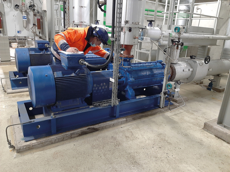 A service worker in an orange work jacket and a blue safety helmet is checking a pump unit in a power station.