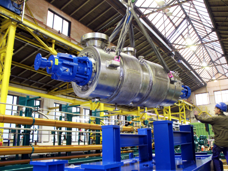 Montage einer schweren Apollo BB5-Hochdruckpumpe (Barrel-Pumpe) mit integriertem Heizmantel in der Produktionshalle; die Pumpe schwebt an einem Industriekran über dem massiven Grundrahmen während der präzisen Positionierung.