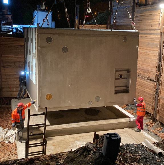 Zu sehen ist eine Baustelle bei Nacht, auf der ein Wassercontainer mithilfe eines Krans von einer Druckerhöhungslage auf ein Betonfundament gesetzt wird. Zwei Arbeiter in leuchtender Arbeitskleidung helfen bei der genauen Positionierung.