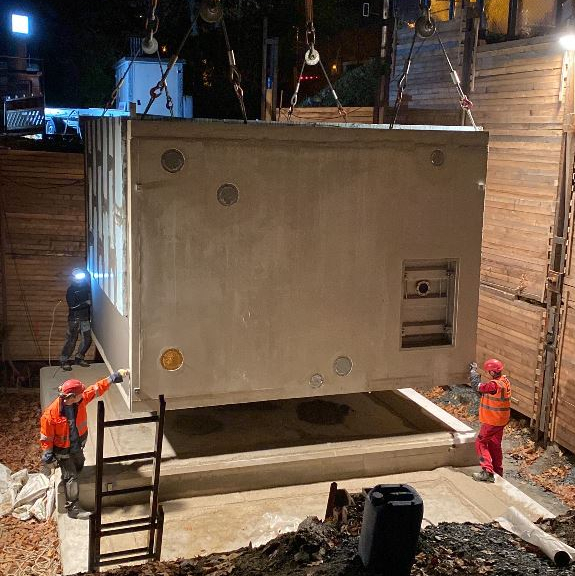 The image shows a construction site at night, where a water container is being placed on a concrete foundation from a pressure booster station using a crane. Two workers in high-visibility clothing are assisting with the precise positioning.