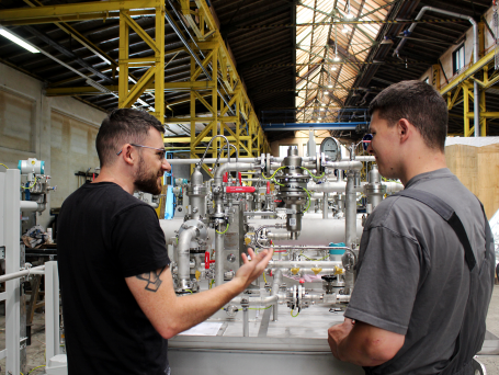 Lachende junge Männer erzählen vor einem Apollo Schmierölsystem. Im Hintergrund ist eine Industriehalleneinrichtung zu sehen.