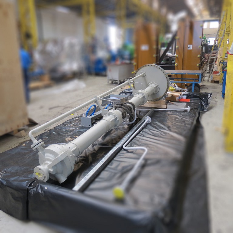 A vertical KRHV pump is placed on a wooden packaging base lined with black foil. Accessories such as pipes and spare parts are located next to the pump. Apollo's production hall can be seen in the background, albeit very blurred.
